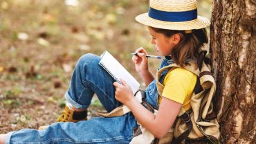 Child sitting outdoors reading a notebook, wearing a straw hat and backpack.