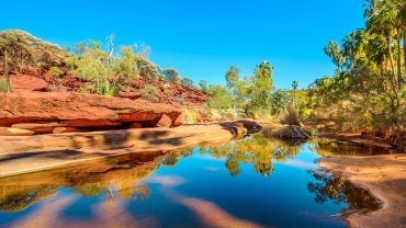 Australian outback river with red rocks and lush greenery under a clear blue sky.