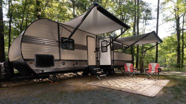 Modern RV trailer with awnings and outdoor seating in a wooded area.