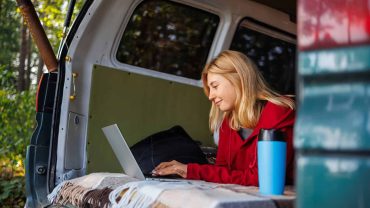 Camping van with woman using laptop outdoors in nature setting.