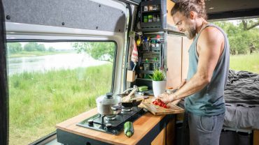 Man cooking in a camper van kitchen with scenic outdoor view.