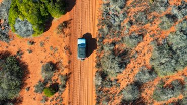 Aerial view of a vehicle driving on a dirt road through Australian outback with sparse trees and red.