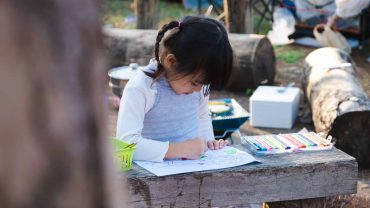 Young girl studying outdoors at a rustic wooden table with books and stationery.