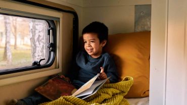 Child reading a book while sitting on a train bed by the window.
