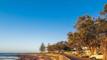 Coastal scene with beach, trees, and parked caravans at sunset.
