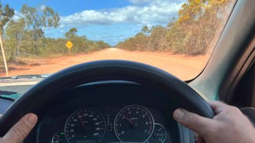 Driving on a rural dirt road with a clear sky and trees in the background.