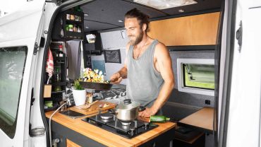 Camping van kitchen with man preparing a meal inside a compact, mobile cooking space.