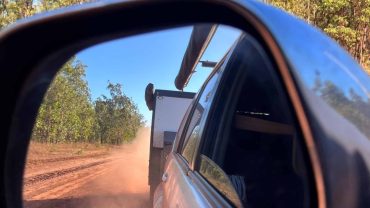 View of a vehicle's side mirror showing a dirt road and trees during a road trip.