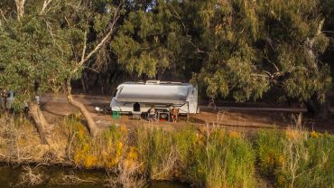 Camper trailer parked near water with trees in the background, outdoor camping scene.